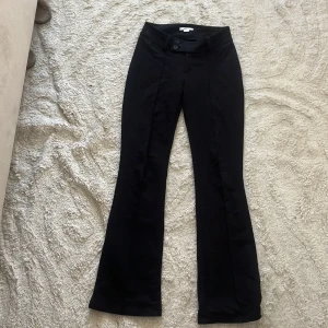 Svarta bootcut kostym byxor från Gina Tricot - Svarta bootcut kostymbyxor från Gina Tricot med  två knappar framtill. Byxorna har pressveck och bälteshällor, vilket ger en clean och stilren look. Perfekta för dig som gillar en klassisk men trendig vibe.