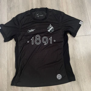 AIK Smokinglirare edition - AIK fotbollströja i svart med diskreta ränder, klubbmärke och årtalet 1891 på bröstet. Nike-logga på framsidan och texten SMOKINGLIRARE på ryggen. Tillverkad i lätt och ventilerande funktionsmaterial, perfekt för match eller träning.