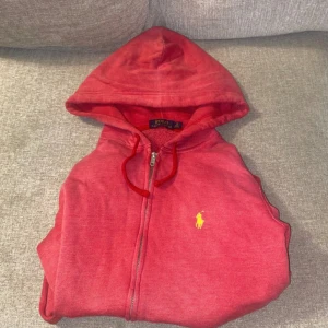 Röd hoodie från Polo Ralph Lauren M - Riktigt snygg röd hoodie från Polo Ralph Lauren i storlek M. Klassisk gul broderad logga på bröstet, dragkedja framtill och huva med snörning. Mjuk insida och skön passform, perfekt för chill dagar. Materialet är bomullsmix.