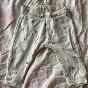 Mönstrade byxor från Newbie, stl 68 - Supersöta ljusa byxor från Newbie i storlek 68, med små blå mönster av luftballonger, flygplan och kameror. Byxorna har resår i midjan och är gjorda i mjuk bomull, perfekta för små äventyrare.