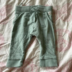 Gröna mjukisbyxor från Lindex Baby - Mysiga ljusgröna mjukisbyxor från Lindex Baby i storlek 56. Byxorna har bred ribbad mudd i midjan och benslut, snörning i midjan och två fickor framtill. Perfekta för små bebisar som vill ha det bekvämt.