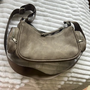 Beige axelväska med dragkedjor - Säljer en beige axelväska i syntetmaterial med bred justerbar rem. Väskan har flera fack med dragkedjor, inklusive två mindre sidofack och ett större huvudfack. Perfekt för att hålla ordning på dina grejer när du är på språng.