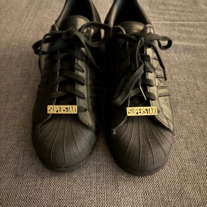 Adidas Superstar Allover Debossed Trefoils - Säljer ett par helsvarta Adidas Superstar sneakers i skinn med klassisk snäcktå och guldiga 'SUPERSTAR'-detaljer på snörningen. Ikoniska tre ränder på sidan och platt sula. (Skorna skall följa med i lådan och med två extra skosnöre också!). Skriv för fler frågor eller bilder i chatten tack!