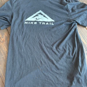 Svart Nike Trail Dri-Fit t-shirt - Svart t-shirt från Nike med Nike Trail-logga på bröstet. Tillverkad i Dri-Fit-material som andas och håller dig torr. Klassisk passform med korta ärmar, perfekt för aktiva dagar eller chill vardagsstil.