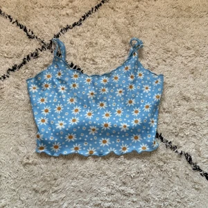 Blommig blå croppad linnetopp SHEIN - Supersöt ljusblå croppad linnetopp från SHEIN med vita och gula prästkragar över hela tyget. Toppen har smala axelband och vågig kant nertill för en extra gullig touch. Perfekt för varma dagar och somriga vibes!