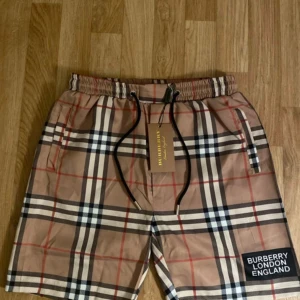 Burberry london England Short  - High Quality  Burberry badshorts i den klassiska beige rutiga designen. Lätta och bekväma med justerbar midja och snörning för perfekt passform. Har “BURBERRY LONDON ENGLAND” logotyp tryckt på benet.