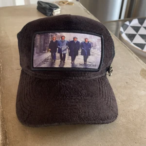 Svart keps från Maggiore med mesh - Svart keps från Maggiore med mjuk manchesterliknande front och mesh baktill. Framsidan har en stor patch med bildmotiv på fyra personer och diskret Maggiore-logga. Sidorna har en silverfärgad M-detalj och justerbar snapback. Perfekt för en cool streetstil.