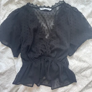 Svart blus med spets från Zara - Supersöt svart blus från Zara med v-ringning och spetsdetaljer längs halsen. Blusen har korta ärmar, prickigt mönster i tyget och volang nertill. Den är lätt transparent och har en feminin, flowy passform som ger en romantisk vibe.