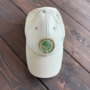 Beige keps med FN-märke - Snygg beige keps med broderat FN-märke framtill i grönt och guld. Märket visar en duva och årtalen 1979-1999. Kepsen är i bomull och har klassisk böjd skärm. Perfekt för dig som gillar unik och stilren accessoar.