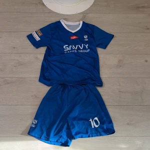 Al Hilal Neymar Jr blå fotbollströja - Blå Al Hilal fotbollströja med Neymar Jr och nummer 10 på ryggen. Kortärmad modell med vit krage och flera sponsorer tryckta på ärmar och bröst. Tillverkad i lätt och ventilerande material, perfekt för match eller träning.