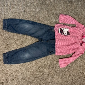 Hello Kitty Set - Skjorta och Jeans - Ett sött set med en Hello Kitty-skjorta med hjärtmönster och ett par blå jeans. Skjortan har långa ärmar, krage och knappar med Hello Kitty-motiv. Jeans är av en ledig modell med resår i midjan och muddar nedtill.