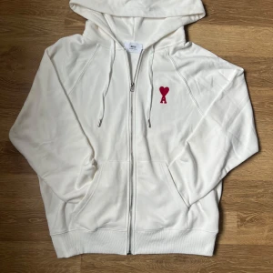 Vit hoodie med röd Ami Paris logo  - Vit hoodie från Ami Paris med dragkedja. På bröstet finns en röd broderad hjärtlogo med bokstaven A. Tröjan har känguruficka och diskret Ami-broderi på ryggen. Tillverkad i mjuk bomull för en clean och stilren look. Sitter väldigt bra och pris är inte fast, snabb leverans. storlek L