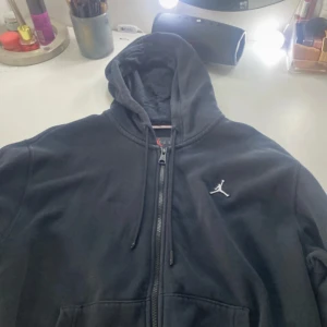 Svart Jordan hoodie med dragkedja - Svart hoodie från Jordan med vit broderad Jumpman-logga på bröstet. Tröjan har hel dragkedja, snörning i huvan och känguruficka. Perfekt för en sportig och avslappnad stil. Pris kan diskuteras!