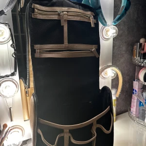 Beige och svart necessärväska - Snygg necessärväska i beige och svart med flera fack och dragkedjor. Perfekt för att organisera smink, borstar eller andra småprylar. Väskan har ett praktiskt handtag upptill och är tillverkad i syntetmaterial med mesh-detaljer på insidan.