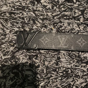 Svart  monogram bälte - Snyggt svart bälte från  med klassiskt monogrammönster och stor logga i metall framtill. Bältet är gjort i skinn med beige insida och svart spänne där är ingraverat. Perfekt accessoar för att lyfta din outfit.