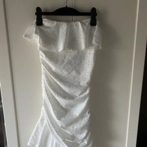 Vit offshoulder kortklänning med volang - Superfin vit kortklänning i bomull med offshoulder och volang upptill. Klänningen har broderade hål, rynkade detaljer och en volangkant nertill. Perfekt för sommarens alla tillfällen och ger en fräsch och trendig look.