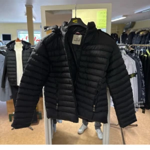 Svart Moncler dunjacka herr - Snygg svart dunjacka från Moncler med klassisk quiltad design och dragkedjor på fickorna. Jackan har lång ärm, hög krage och ikonisk Moncler-logga på ärmen. Perfekt för kalla dagar och ger en clean, stilren look.