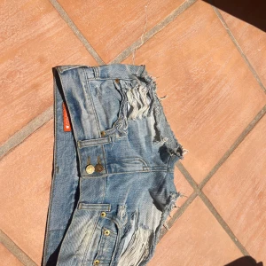 Lowwaist shorts - Säljer ett par blå jeansshorts med råa kanter och slitna detaljer framtill och baktill. Shortsen har klassiska fickor, bälteshällor och guldiga nitar. Perfekta för en avslappnad och trendig look i sommar.