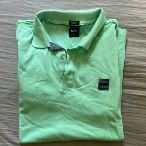 Mintgrön pikétröja från BOSS - Fräsch mintgrön pikétröja från BOSS med klassisk krage, knäppning med två knappar och diskret BOSS-logga på bröstet. Tröjan är i bomull och har korta ärmar, perfekt för en clean och stilren look. Passar även S. Nyskick