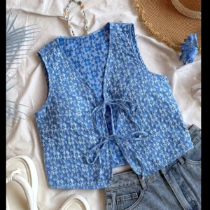 Blå blommig väst med knyt - Supersöt blå väst med ljusblått blommönster och v-ringning. Västen har två knytband framtill och är lätt croppad. Perfekt att styla över en t-shirt eller klänning för en chill sommarlook.