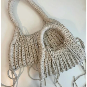 Beige stickad axelväska med handtag - Säljer en beige stickad axelväska med grova handtag och coola knytband längs sidorna. Väskan har en unik, handgjord känsla och passar perfekt för dig som gillar boho-stil. Rymmer det viktigaste och är superenkel att slänga över axeln.