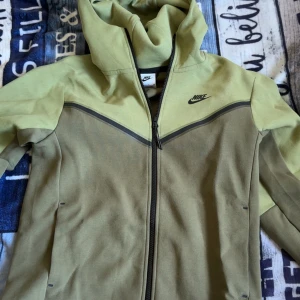 Nike hoodie jacka i olivgrönt - Nike hoodie jacka i olivgrönt med dragkedja och huva. Snygg tvåtonad design med mörkare och ljusare partier, samt svart Nike-logga på bröstet. Jackan har långa ärmar och är tillverkad i mjukt bomullsmaterial, perfekt för chill eller träning. kan säljas endast byxor eller jackan. priset ej hugget i sten vid snabbaffär 