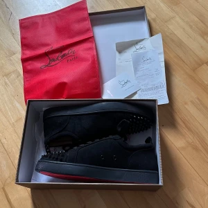 Christian Louboutin svarta sneakers med nitar - Svarta sneakers från Christian Louboutin i mocka med ikoniska röda sulor. Skorna har svarta nitar på tån, snörning och låg profil. Snygg och edgy design som sticker ut, perfekt för dig som gillar statement-detaljer.