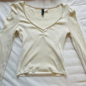 Ribbad vit långärmad topp - Supersöt ribbad långärmad topp i vit färg. Djup v-ringning med spetskant och rynkdetalj framtill. 