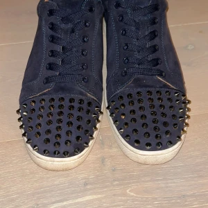 Christian Louboutin  - Mörkblå sneakers från Christian Louboutin i mocka med svarta nitar på tån och ikonisk röd sula. Skorna har snörning och vit platt sula. Kommer med originalkartong och dustbag. Perfekta för dig som vill sticka ut med en edgy vibe.