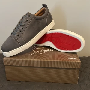 Grå sneakers från Christian Louboutin - Snygga grå sneakers från Christian Louboutin i mocka med klassisk röd sula och vita kanter. Skorna har rund tå, platt sula och snörning. Insidan är fodrad i beige läder för extra komfort. Perfekta för dig som vill ha en stilren och lyxig look.