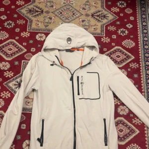 Vit vindjacka från Sail Racing - Snygg vit vindjacka från Sail Racing med huva och svarta detaljer. Jackan har dragkedjor på fickorna och en cool logga på bröstet samt på huvan. Perfekt för blåsiga dagar och har en sportig vibe med orange insida vid dragkedjan.