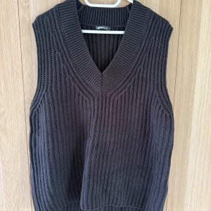 Svart stickad väst från Gina Tricot - Säljer en svart, grovstickad väst från Gina Tricot i storlek XS. Västen har en djup v-ringning och bred ribbad kant runt halsen. Perfekt att slänga över en skjorta eller t-shirt för en chill och trendig look.