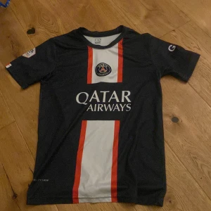 PSG Neymar Jr 10 matchtröja - Snygg PSG fotbollströja med Neymar Jr och nummer 10 på ryggen. Tröjan är mörkblå med en vit och röd rand längs mitten, PSG-logga på bröstet och Qatar Airways som sponsor. Tillverkad i polyester, kortärmad och perfekt för dig som älskar fotboll och Neymar.