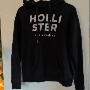 Svart Hollister hoodie med tryck - Svart hoodie från Hollister med stor vit broderad logga på bröstet och texten 'CALIFORNIA' längs ena ärmen. Klassisk känguruficka, huva med snörning och mjukt material. Perfekt för en chill och avslappnad stil.