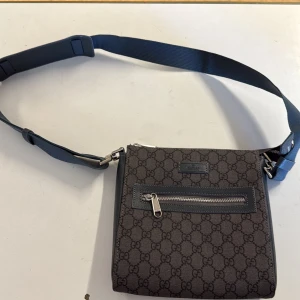 Gucci axelväska i mörkgrå canvas - Snygg axelväska från Gucci i mörkgrå canvas med klassiskt GG-mönster. Väskan har justerbar axelrem i blått, detaljer i skinn och silverfärgade dragkedjor. Perfekt storlek för det viktigaste och en stilren look med lyxig känsla.