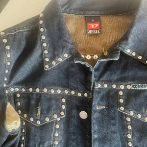 Diesel jeansjacka med nitar, mörkblå - Snygg mörkblå jeansjacka från Diesel med  silverfärgade nitar längs sömmarna och fickorna. Klassisk krage, knäppning framtill och två bröstfickor. Jackan har detaljer i satin vid armhålorna som ger en unik touch. Perfekt för dig som vill sticka ut.  Från 1998