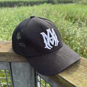 Svart keps från Amiri - Svart truckerkeps från Amiri med vit broderad logga framtill. Kepsen har mesh på sidorna och bak, samt justerbar snapback för perfekt passform. Klassisk böjd skärm och cool streetkänsla – perfekt för dig som gillar exklusiva accessoarer.