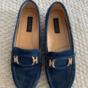 Mörkblå mockaloafers från Novita - Snygga mörkblå loafers i mocka från Novita med guldfärgade metalldetaljer framtill. Skorna har en klassisk rund tå och platt sula med gummidobbar för extra grepp. Säljer pga köpte för små. Nypris 1450kr.