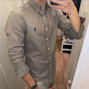 Grå slim fit skjorta från Ralph Lauren - Snygg grå skjorta från Ralph Lauren i slim fit-modell. Klassisk button-down krage, långa ärmar och broderad marinblå logga på bröstet. Skjortan är i bomull och har vita knappar framtill och vid ärmsluten. Perfekt för en clean och stilren look.