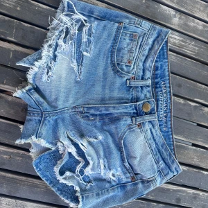 Blå  jeansshorts American Eagle - Snygga blå jeansshorts från American Eagle med slitna detaljer, fransiga kanter och råa avslut. Klassisk femficksmodell med knapp och dragkedja. Extra cool detalj med spets under slitningarna framtill. Perfekta för sommaren! Köptes här på Plick för ca 300, använda två gånger. Dom är typ mid rise till low rise 
