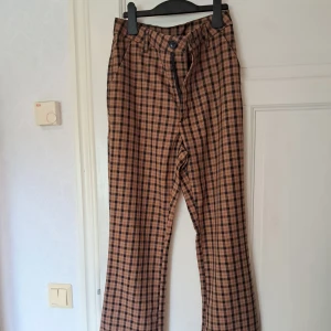 Coola rutiga bootcut byxor från H&M  - Snygga rutiga bootcut byxor från H&M i brun och svart. Vida ben och normal passform, tillverkade i mjuk polyester med lite stretch. Benen har slits nertill i sidorna se sista bilden. Perfekta för dig som gillar en rockig och alternativ look. Stängs med knapp och dragkedja framtill.