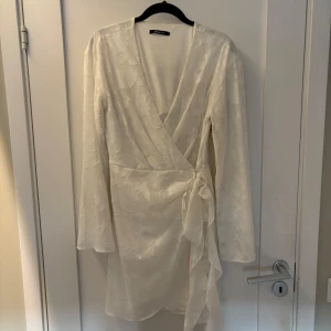 Vit omlottklänning med knytdetalj - Vit kort omlottklänning från Gina Tricot med blommönster i tyget. Klänningen har långa ärmar, v-ringning och knyt i midjan. Endast använd någon gång.