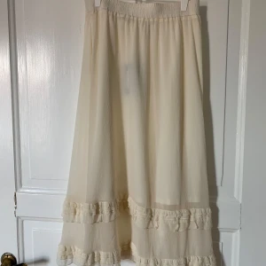 Krämvit långkjol med volangdetaljer - Superfin långkjol i krämvitt med volangdetaljer nertill. Kjolen har en luftig och flowy känsla med resår i midjan för extra komfort. Perfekt för dig som gillar en romantisk och bohemisk stil. Passar lika bra till sneakers som sandaler.