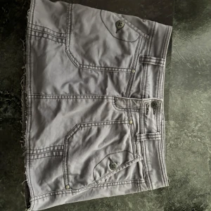 Grå jeanskjol med rå kant - Säljer en grå kort jeanskjol med rå, fransig kant nertill. Kjolen har stora fickor både fram och bak med coola knappar och bälteshällor i midjan. Perfekt för dig som gillar en avslappnad och trendig stil. (INTE 32 kjol storlek men 32 tum väldigt stor skillnad)