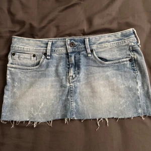 Blå jeanskjol från Pepe Jeans - Snygg ljusblå jeanskjol från Pepe Jeans med slitna detaljer och rå kant nertill. Klassisk femficksmodell med bälteshällor och knappstängning. Perfekt för dig som gillar en avslappnad och trendig stil.