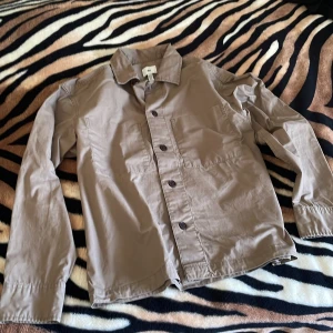 Beige overshirt från H&M - Snygg beige overshirt från H&M med klassisk krage och knäppning framtill. Jackan har raka linjer, stora fickor på bröstet och är tillverkad i bomull. Perfekt att slänga över en hoodie eller t-shirt för en chill look.