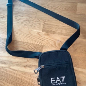 Svart axelväska från EA7 Emporio Armani - Snygg svart axelväska från EA7 Emporio Armani med justerbart band och dragkedja framtill. Perfekt storlek för mobil, plånbok och andra småsaker. Stilren design med vit logga på framsidan och praktiskt ytterfack.