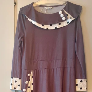 *NY*Lila klänning med prickiga detaljer i stl L  - Unik lila klänning med långa ärmar och vita detaljer med svarta prickar vid krage, ärmslut och höft. Dekorativa vita knappar framtill ger en retrovibe. Mjuk och skön, perfekt för dig som gillar lekfull stil.