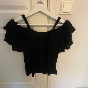 Svart volangig blus med axelband - Snygg svart blus med volanger och tunna axelband. Offshoulder-stil med dubbla lager volanger som ger en luftig och trendig look. Perfekt för dig som gillar en feminin och cool vibe. Tunn och lätt i materialet.