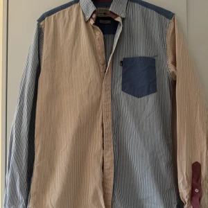 Randig skjorta från Lexington - Snygg skjorta från Lexington med smala ränder i blått och beige, kontrasterande blå bröstficka och axelparti samt vinröda detaljer på insidan av kragen och ärmsluten. Klassisk krage, långärmad och knäppning framtill. Perfekt för dig som gillar unika detaljer.